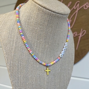 May include: A pastel rainbow beaded necklace with a gold cross charm and the name "Fiona" spelled out in white beads.