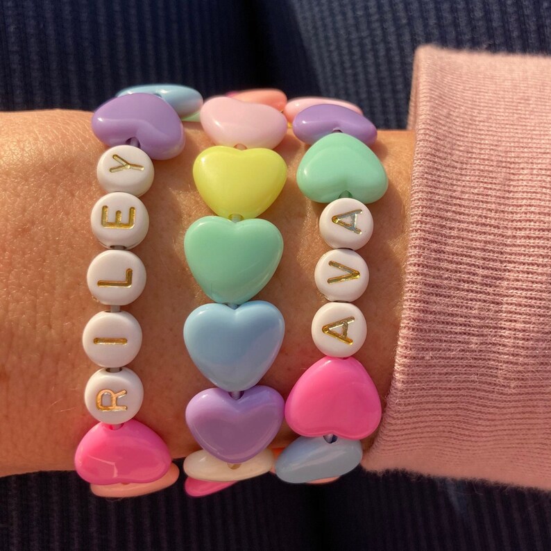 May include: Three heart-shaped beaded bracelets with the names Riley, Ava, and Y spelled out in gold letters on white beads.