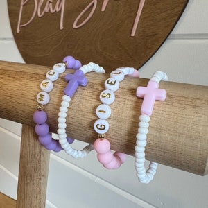 May include: Four bracelets with cross charms and personalized names. The bracelets are made of white and purple beads with gold lettering. The names on the bracelets are "MAC", "GISELLE", and two other names that are not visible in the image.