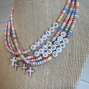 May include: Multi-layered pastel beaded necklace with white beads that spell out the names "Adelyn", "Myla", "Jordyn", and "Mina". Each layer has a small gold cross charm.