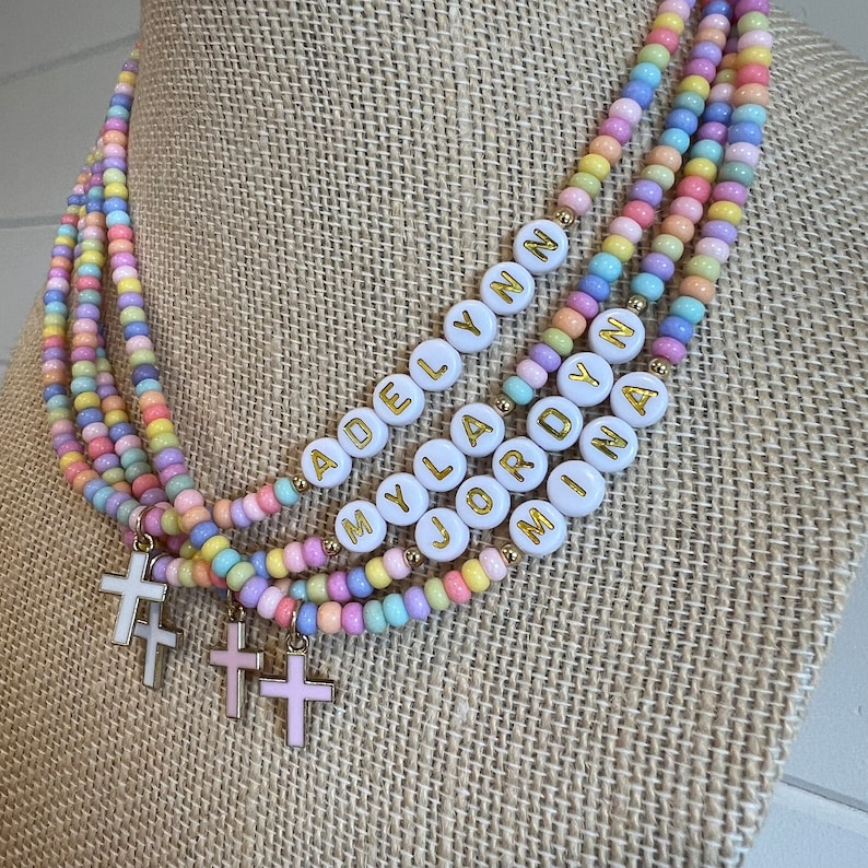 May include: Multi-layered pastel beaded necklace with white beads that spell out the names "Adelyn", "Myla", "Jordyn", and "Mina". Each layer has a small gold cross charm.