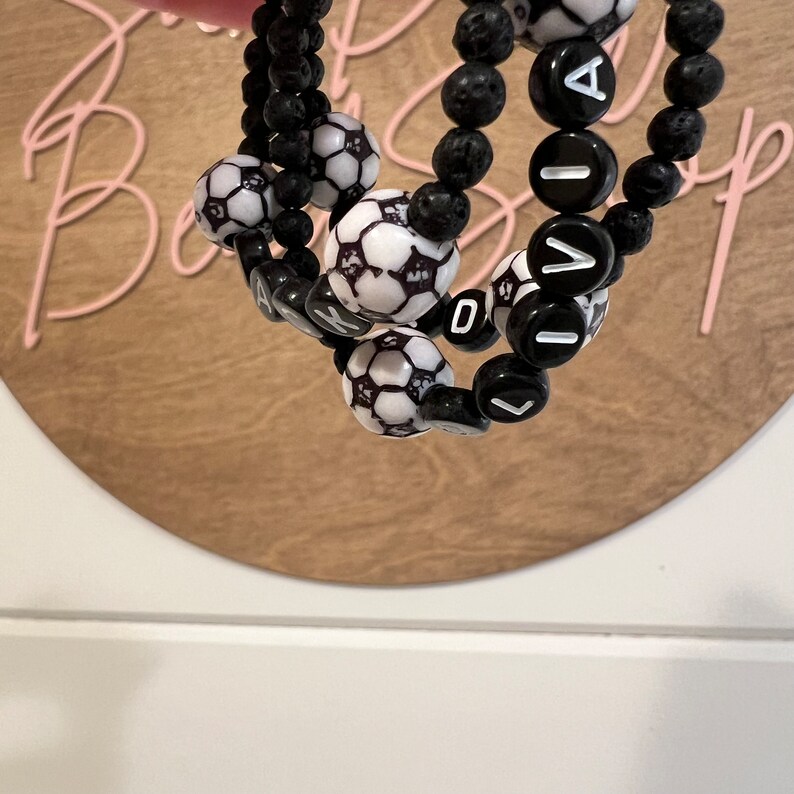 May include: Three black lava rock bracelets with white soccer ball beads and black letter beads spelling "LIVIA".