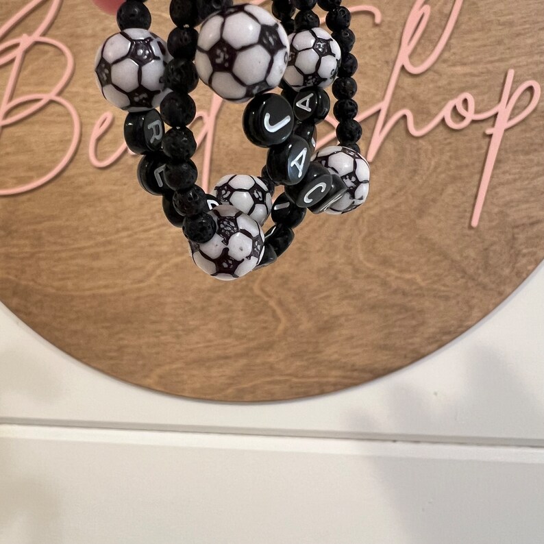 K&ouml;nnte beinhalten: Schwarze Lava-Stein-Armb&auml;nder mit wei&szlig;en Fu&szlig;ball-Perlen und schwarzen Buchstabenperlen, die den Namen "JACK" buchstabieren.