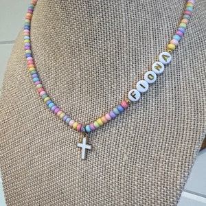May include: A pastel-colored beaded necklace with a gold cross charm and the name "Fiona" spelled out in white beads.