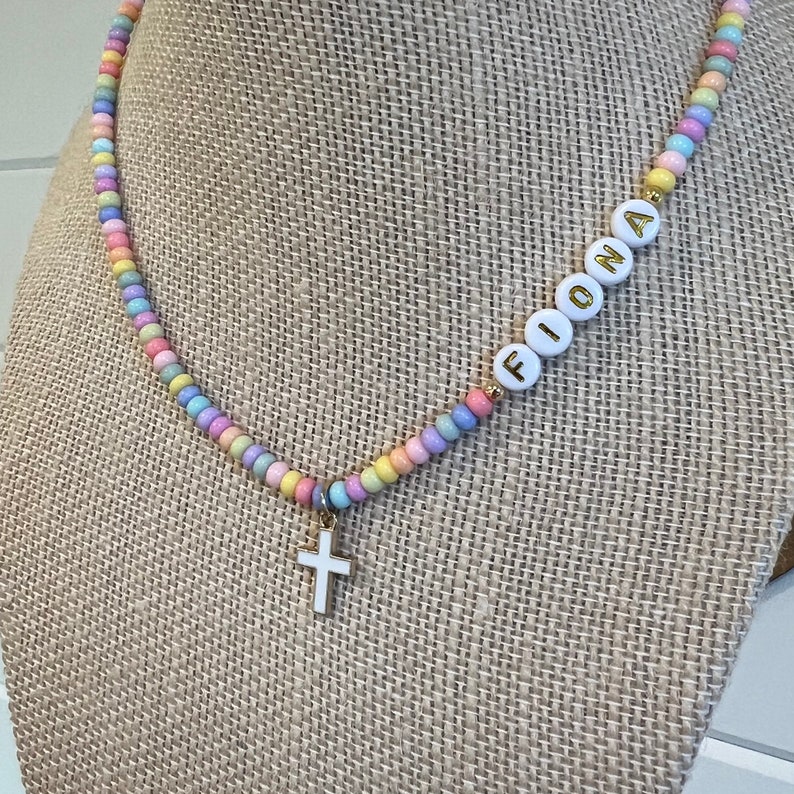 May include: A pastel-colored beaded necklace with a gold cross charm and the name "Fiona" spelled out in white beads.