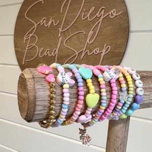 May include: A collection of colorful beaded bracelets with various charms, including hearts, cats, stars, and letters. The bracelets are displayed on a wooden stand. San Diego Bead Shop is written on a wooden sign in the background.