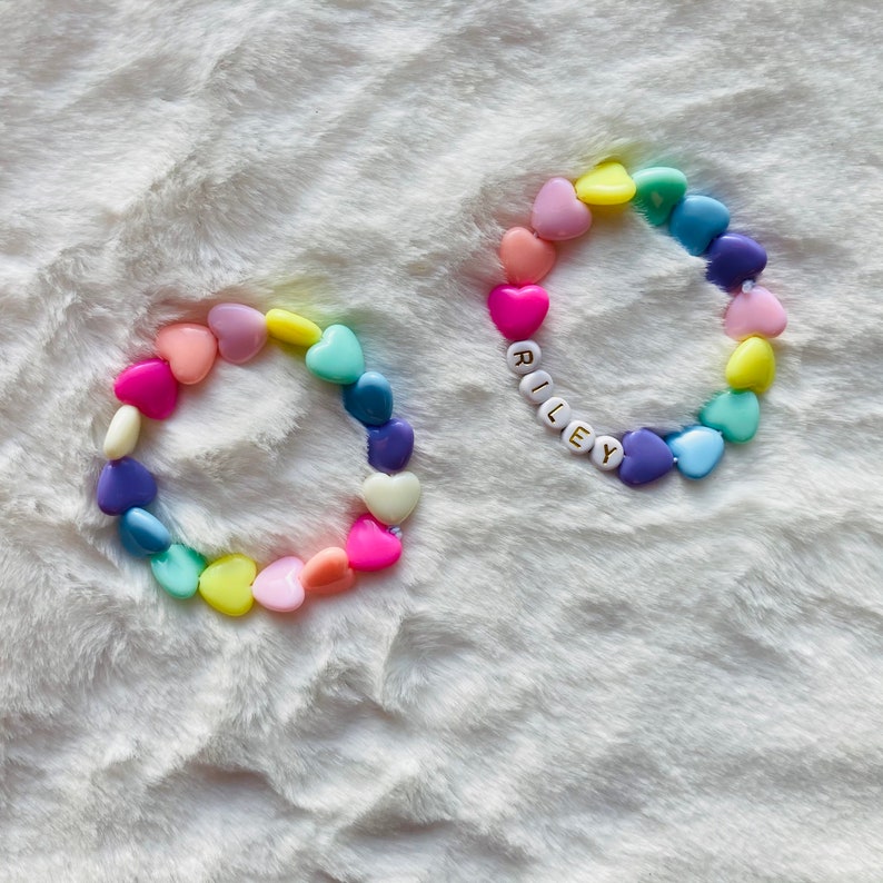 May include: Two colorful heart-shaped bead bracelets. One bracelet spells out the name "Riley" in gold letters on white beads. Both bracelets are on a white fuzzy surface.