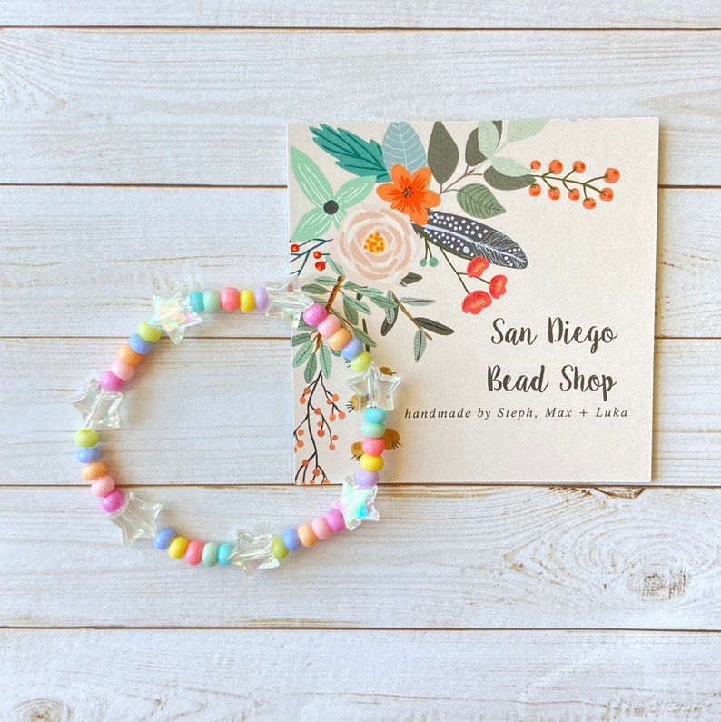 May include: A colorful beaded bracelet with iridescent star-shaped charms. The bracelet is made with pastel pink, blue, yellow, and green beads. The bracelet is displayed on a wooden surface with a business card that reads "San Diego Bead Shop, handmade by Steph, Max + Luka".