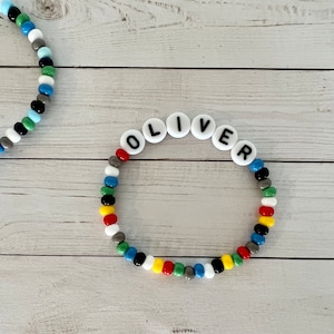 以下が含まれることがあります： 黒文字で「OLIVER」と綴られた白いビーズでできた、カラフルなビーズブレスレットです。