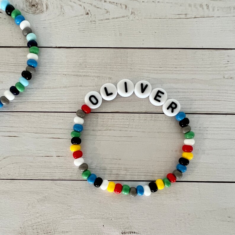 以下が含まれることがあります： 黒文字で「OLIVER」と綴られた白いビーズでできた、カラフルなビーズブレスレットです。