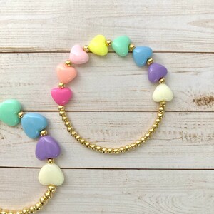 Puede incluir: Dos pulseras de oro con cuentas en forma de coraz&oacute;n y dijes de coraz&oacute;n de colores pastel. Las pulseras est&aacute;n sobre un fondo de madera blanco.