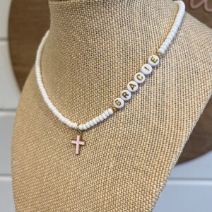 May include: White beaded necklace with a gold cross charm and the name "Gracie" spelled out in gold letters on the beads.
