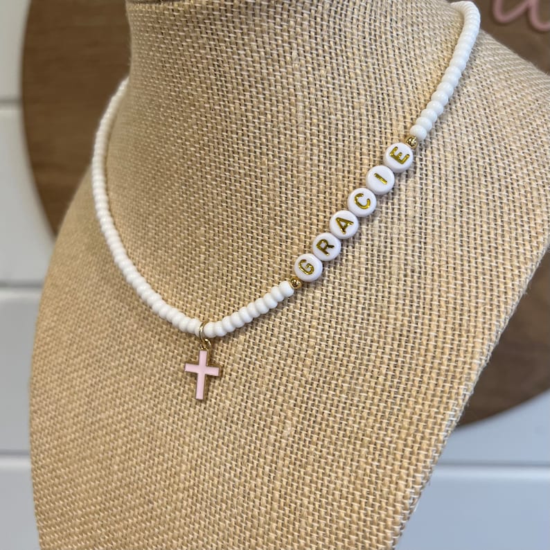 May include: White beaded necklace with a gold cross charm and the name "Gracie" spelled out in gold letters on the beads.