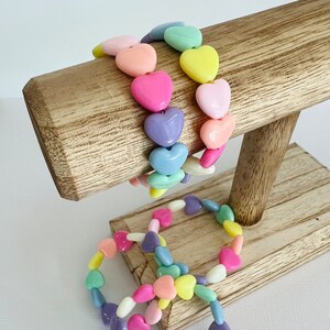 May include: Three bracelets made of pastel colored heart-shaped beads. The bracelets are displayed on a wooden stand.