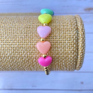 Puede incluir: Un brazalete de corazones arco&iacute;ris con cuentas doradas. El brazalete presenta cinco cuentas en forma de coraz&oacute;n en tonos de verde, amarillo, morado, rosa y rojo.