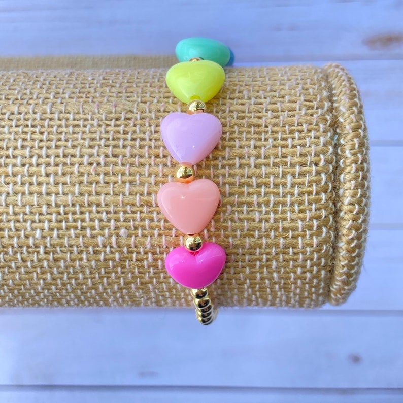 Puede incluir: Un brazalete de corazones arco&iacute;ris con cuentas doradas. El brazalete presenta cinco cuentas en forma de coraz&oacute;n en tonos de verde, amarillo, morado, rosa y rojo.