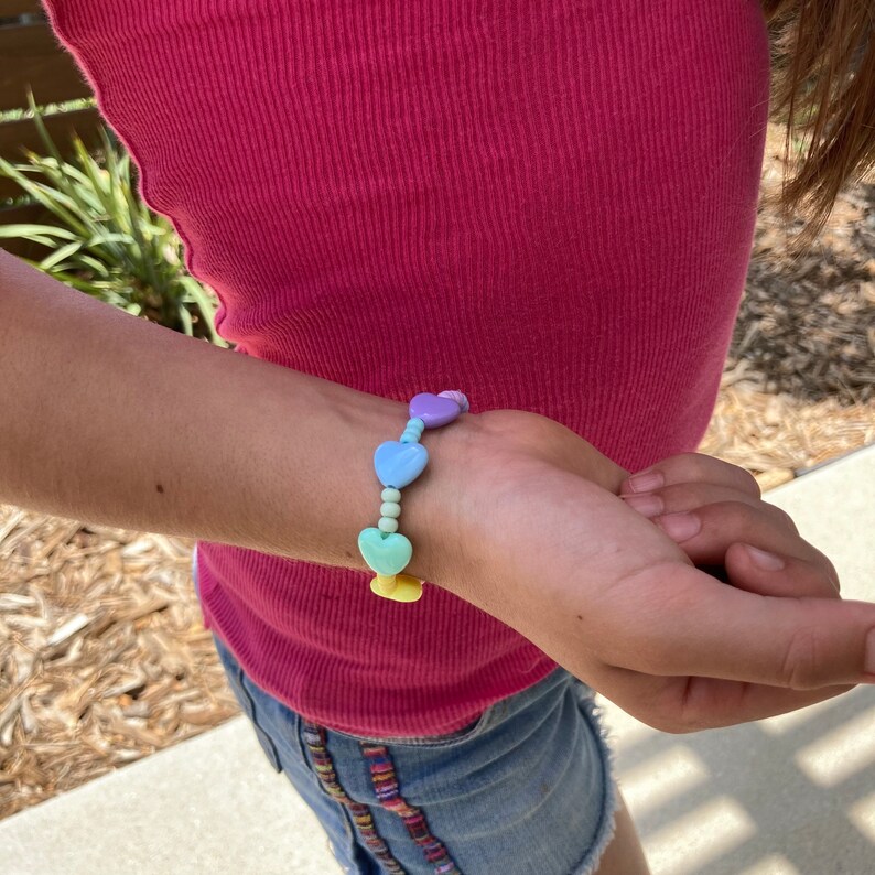 May include: A colorful heart-shaped beaded bracelet with pastel blue, purple, green, and yellow beads. The bracelet is worn on a person's wrist.