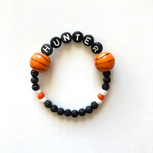 May include: Black and orange beaded bracelet with the name "HUNTER" spelled out in white letters. The bracelet also features two orange basketball-shaped beads.
