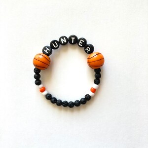 K&ouml;nnte beinhalten: Schwarzes und orangefarbenes Armband mit Basketball-Motiv, auf dem der Name "HUNTER" in wei&szlig;en Buchstaben auf schwarzen Perlen geschrieben ist.