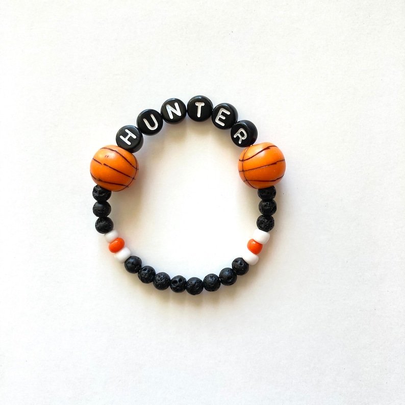 K&ouml;nnte beinhalten: Schwarzes und orangefarbenes Armband mit Basketball-Motiv, auf dem der Name "HUNTER" in wei&szlig;en Buchstaben auf schwarzen Perlen geschrieben ist.