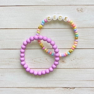 May include: Two bracelets, one with pastel colored beads and the name "Cece" spelled out in white beads with gold lettering, and the other a solid purple bracelet.