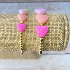 Puede incluir: Dos pulseras de cuentas doradas con cuentas en forma de coraz&oacute;n en tonos rosa, melocot&oacute;n y lavanda.