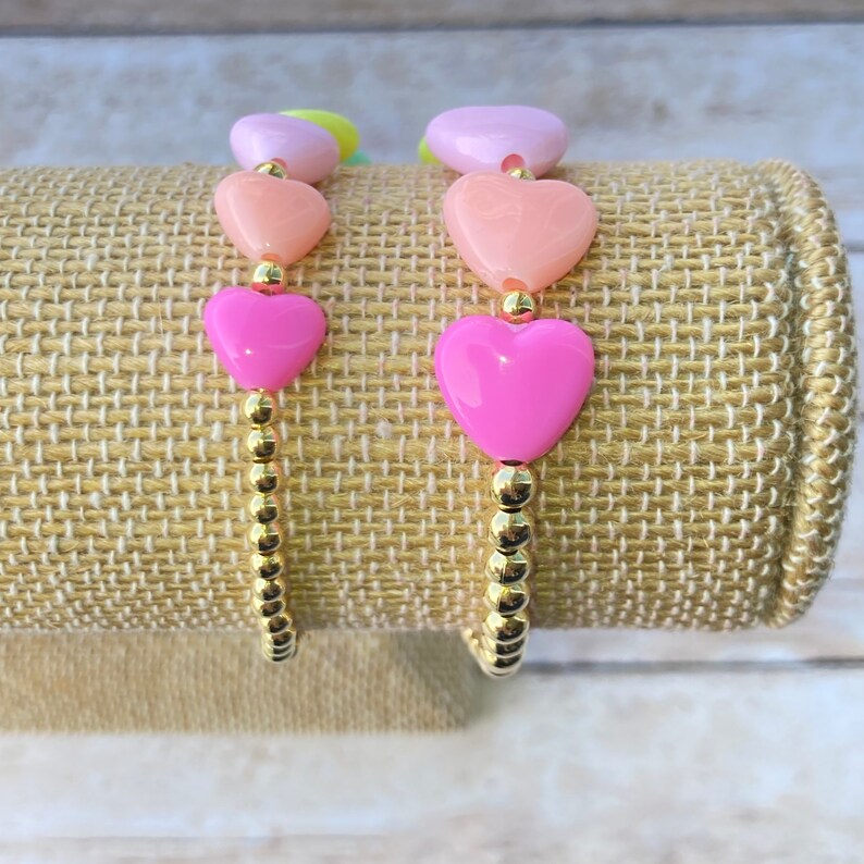 Puede incluir: Dos pulseras de cuentas doradas con cuentas en forma de coraz&oacute;n en tonos rosa, melocot&oacute;n y lavanda.