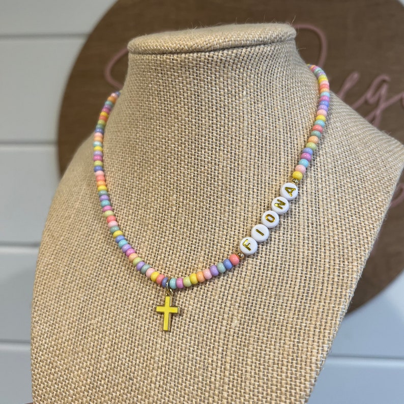 May include: A pastel-colored beaded necklace with a gold cross charm and the name "Fiona" spelled out in white beads.
