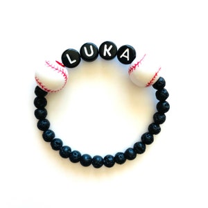 K&ouml;nnte beinhalten: Armband aus schwarzen Lavasteinen mit wei&szlig;en Baseball-Anh&auml;ngern und schwarzen Buchstabenperlen, die "LUKA" buchstabieren.