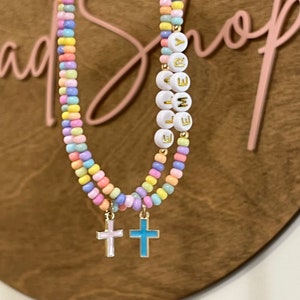 May include: A pastel-colored beaded necklace with a charm of a gold cross. The necklace has white beads with gold letters spelling the name "EMILY".