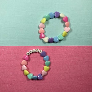 May include: Two heart-shaped bead bracelets. The top bracelet is a rainbow of colors and the bottom bracelet spells out the name "RILEY" in gold letters.