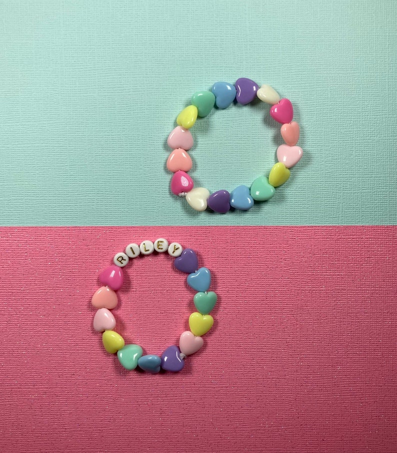 May include: Two heart-shaped bead bracelets. The top bracelet is a rainbow of colors and the bottom bracelet spells out the name "RILEY" in gold letters.