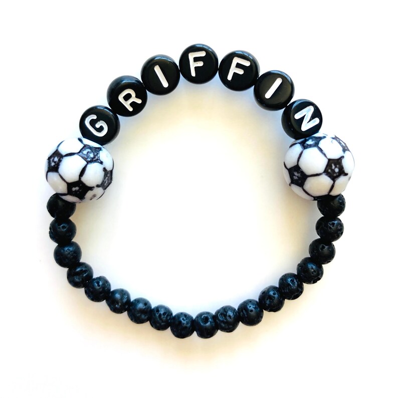 May include: Black beaded bracelet with the name "GRIFFIN" spelled out in white letters. The bracelet has two black and white soccer ball charms.