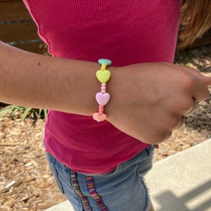 May include: A colorful heart-shaped beaded bracelet with pastel pink, yellow, and green beads. The bracelet is on a person's wrist.