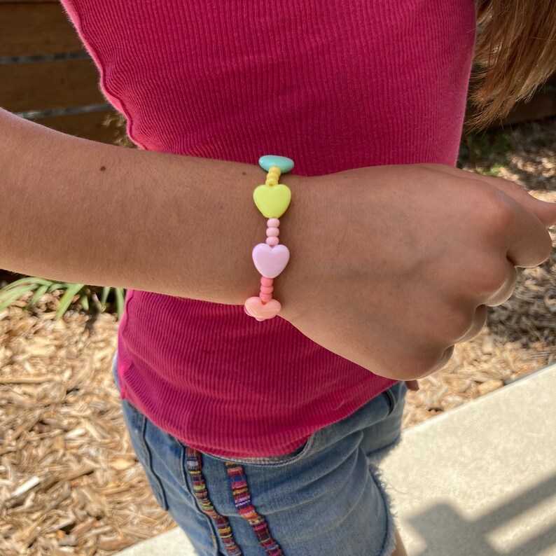 May include: A colorful heart-shaped beaded bracelet with pastel pink, yellow, and green beads. The bracelet is on a person's wrist.