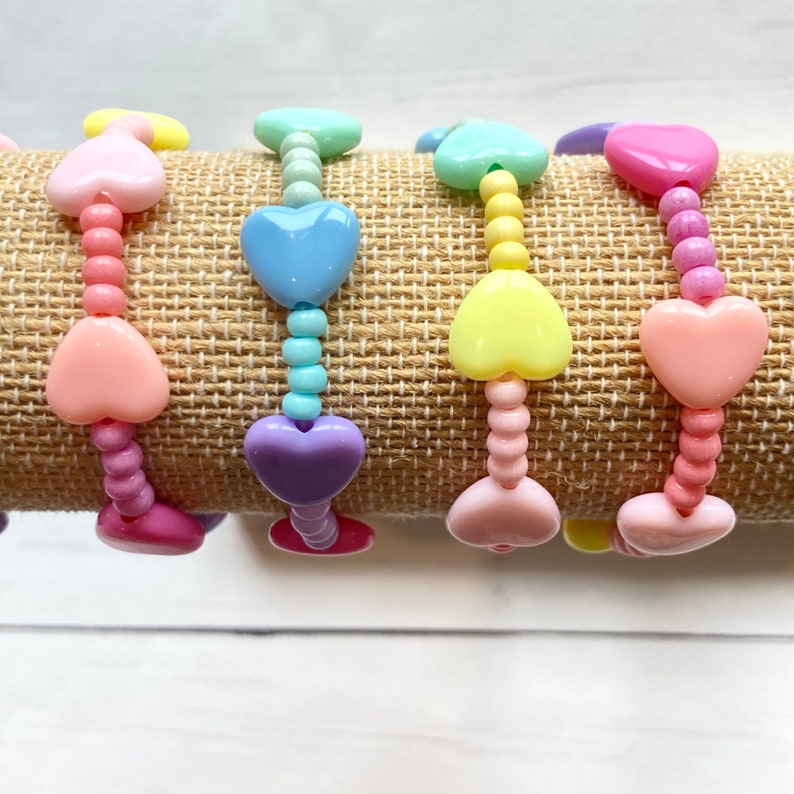 May include: Five colorful heart-shaped beaded bracelets in pastel pink, light blue, purple, yellow, and pink. The bracelets are stacked on a brown burlap surface.