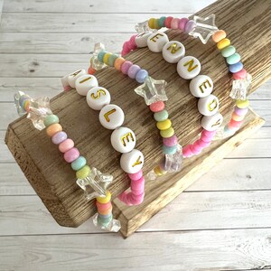 May include: Four colorful beaded bracelets with star charms and white beads with gold lettering spelling out the names "Ainsley", "Kennedy", "En", and "Ley".