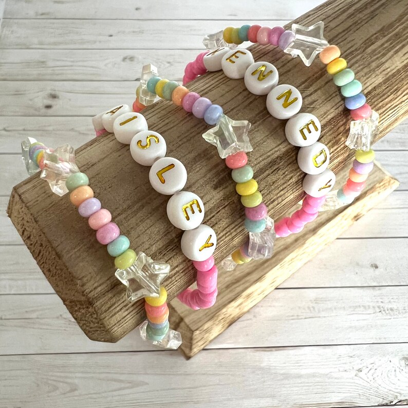 May include: Four colorful beaded bracelets with star charms and white beads with gold lettering spelling out the names "Ainsley", "Kennedy", "En", and "Ley".