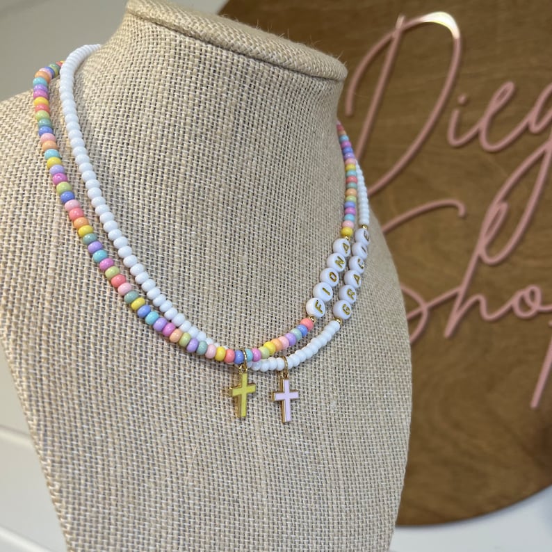 May include: A colorful beaded necklace with the names "Fiona" and "Grace" spelled out in white beads. The necklace has two gold cross charms.