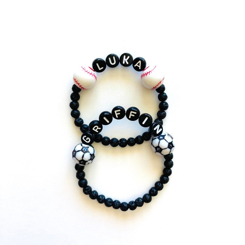 K&ouml;nnte beinhalten: Zwei schwarze Perlenarmb&auml;nder mit den Namen Luka und Griffin in wei&szlig;en Buchstaben. Die Armb&auml;nder sind mit schwarz-wei&szlig;en Fu&szlig;b&auml;llen und wei&szlig;en Baseballs verziert.