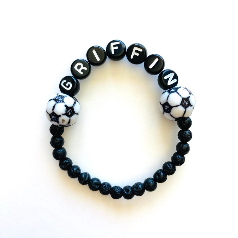 K&ouml;nnte beinhalten: Schwarzes Lava-Armband mit wei&szlig;en Buchstabenperlen, die "GRIFFIN" buchstabieren, und schwarzen und wei&szlig;en Fu&szlig;ballanh&auml;ngern.