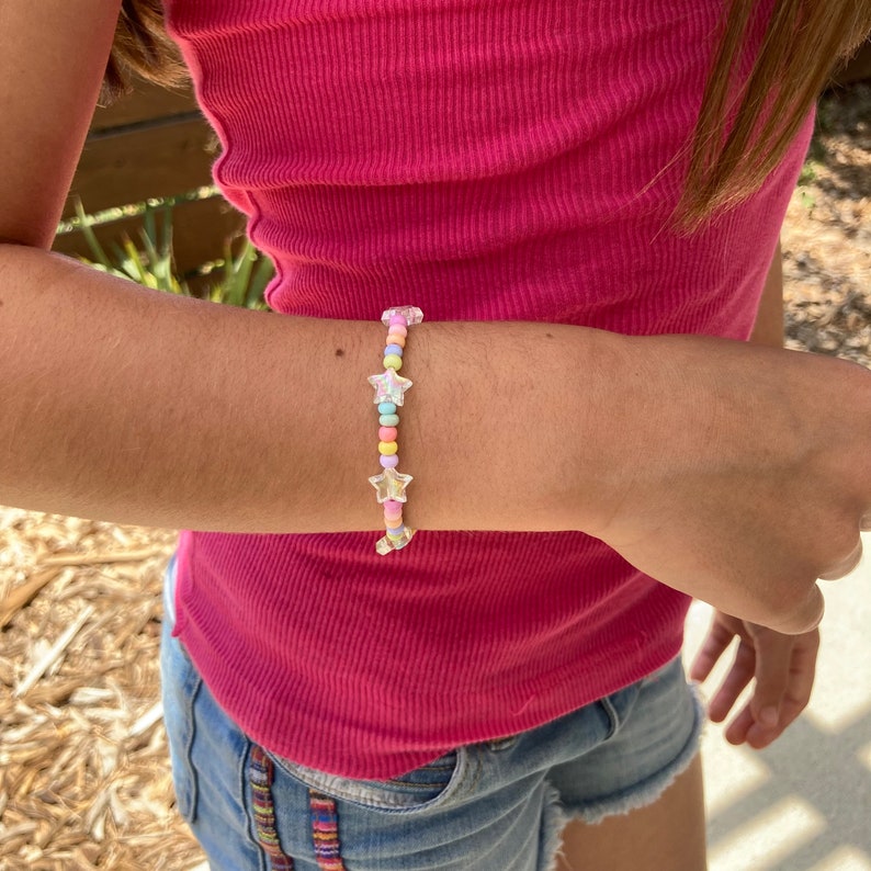 May include: A colorful beaded bracelet with star-shaped charms. The beads are pastel pink, blue, yellow, and green. The bracelet is on a person's wrist.