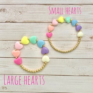 Puede incluir: Dos pulseras con cadena dorada y cuentas de coraz&oacute;n de colores. La pulsera de arriba tiene corazones m&aacute;s peque&ntilde;os y la pulsera de abajo tiene corazones m&aacute;s grandes. El texto "SMALL HEARTS" est&aacute; encima de la pulsera de arriba y el texto "LARGE HEARTS" est&aacute; debajo de la pulsera de abajo.
