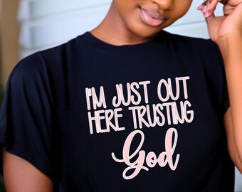 Pode incluir: Camiseta preta com o texto "I'M JUST OUT HERE TRUSTING God" em escrita branca e rosa. A modelo usa uma saia preta e um cinto estampado. O fundo inclui uma parede branca e folhagem verde.