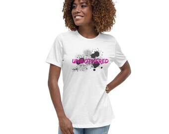 May include: White t-shirt with the word "UNBOTHERED" in pink, with black and gray paint splatter design. The shirt has short sleeves and a relaxed fit. The t-shirt is a casual wear item.