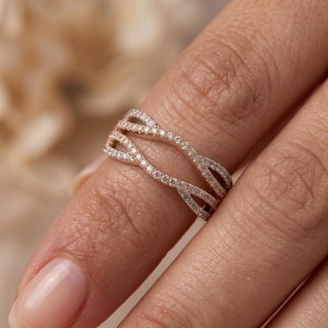 May include: A rose gold ring featuring a bypass design with two overlapping bands. Each band is encrusted with small, clear pavé-set stones that follow the wavy, intertwined silhouette of the metal, creating a continuous sparkling effect.
