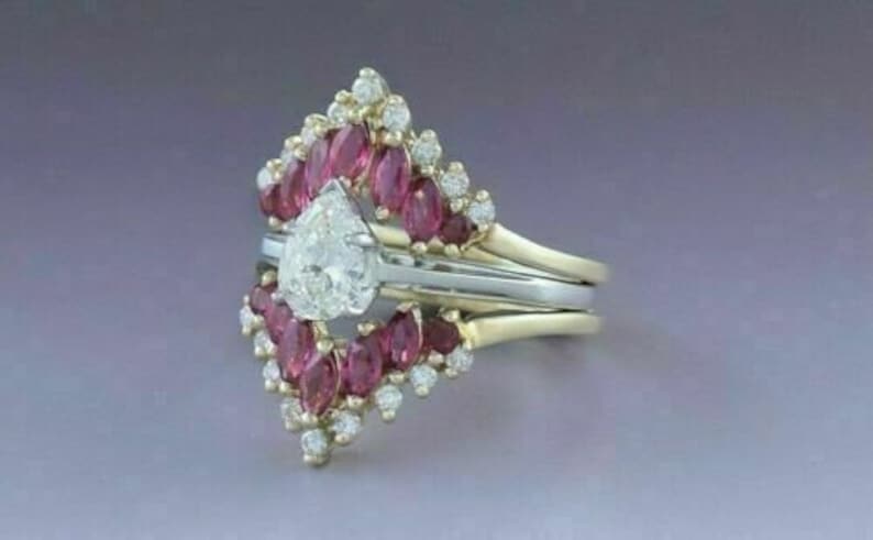 Puede incluir: Un anillo de compromiso de diamante de forma de pera engastado en una banda de oro, rodeado por un halo de diamantes y rub&iacute;es m&aacute;s peque&ntilde;os.