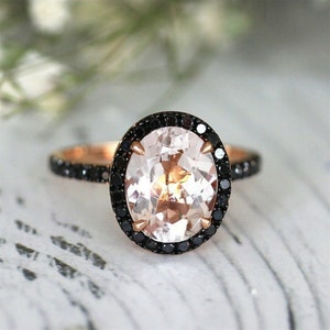 May include: A rose gold engagement ring with a large oval-shaped pink gemstone surrounded by a halo of black diamonds.