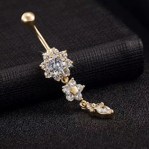 May include: Gold belly button ring with a dangling charm featuring three flower-shaped designs with clear rhinestones.