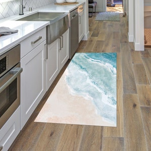 Puede incluir: Una cocina blanca con gabinetes blancos y un horno y un fregadero de acero inoxidable. Una alfombra azul y blanca con temática de playa está en el suelo.